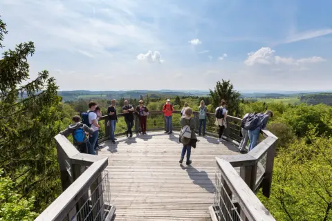 Besucher genießen die Aussicht vom Baumwipfelpfad NRW. Eine Gruppe steht auf der Holzplattform, umgeben von grünen Wäldern und blauen Himmel.