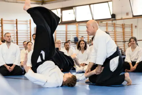 Två personer i vit gi utför en kastteknik inom kampsporten aikido. Åskådare sitter på golvet och observerar.