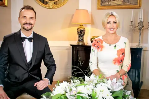 Teresa Werner i jej mąż, elegancko ubrani, siedzą przy bukiecie kwiatów.
