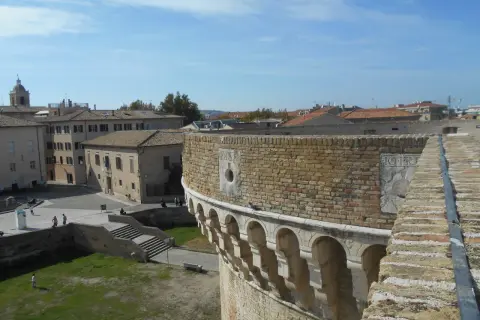 Vista panoramica di Senigallia, Ancona, con un'antica fortezza in primo piano e tetti di tegole rosse sullo sfondo sotto un cielo azzurro.