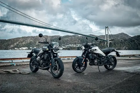 Dwa motocykle Husqvarna Svartpilen 250 stoją nad wodą, pod mostem wiszącym, w malowniczym, górzystym krajobrazie.