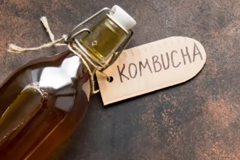 En flaska med en mörk, bubblig kombucha dryck. En brun etikett med texten "Kombucha" hänger på flaskhalsen.