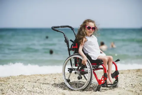 Dziewczynka w wózku inwalidzkim, w białej sukience i okularach przeciwsłonecznych, siedzi na plaży. W tle widać morze i ludzi kąpiących się w wodzie.