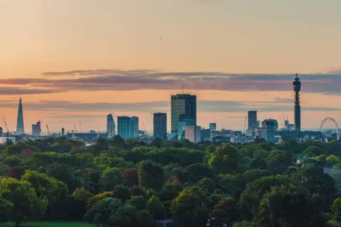 London im Sommer: Die Skyline mit dem Shard und dem BT Tower ragt über einem grünen Park auf, während die Sonne den Himmel in warme Farben taucht.