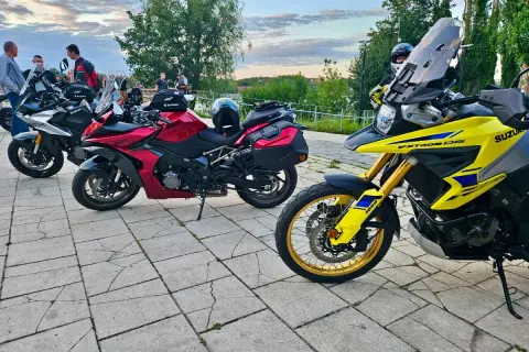 Kilka motocykli Suzuki zaparkowanych na brukowanym placu, z ludźmi w tle.