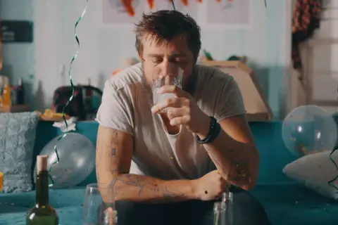 Zmęczony mężczyzna z tatuażami pije wodę po imprezie, symbolizującą jego odstawienie alkoholu. W tle resztki zabawy.