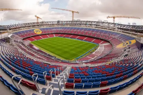 Widok na stadion w budowie, z czerwono-niebieskimi trybunami i zieloną murawą. W tle widoczne są dźwigi, sugerujące, że to jeden z największych stadionów w Polsce.