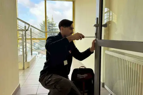 Mężczyzna próbuje otworzyć drzwi bez klucza, używając śrubokręta. Obok niego stoi torba z narzędziami.