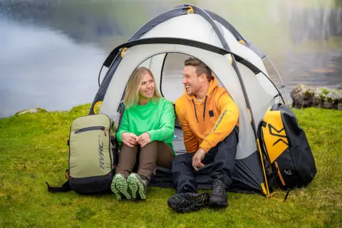 Ett glatt par njuter av att campa i skogen vid en sjö. De sitter i sitt tält med ryggsäckar bredvid.