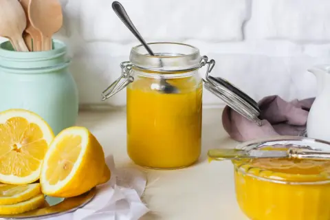 Vasetto di vetro pieno di lemon curd, con limoni freschi affettati e un cucchiaino.