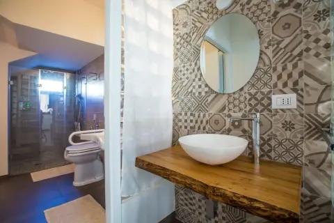 Bagno piccolo con antibagno, doccia in vetro, WC e lavabo su mensola in legno grezzo. Piastrelle geometriche e specchio rotondo.