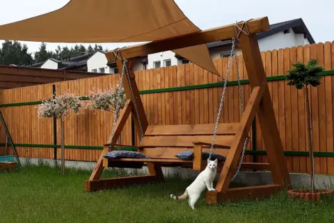 Drewniana huśtawka w ogrodzie, z beżowym daszkiem i poduszkami. Biały kotek przygląda się aranżacji.