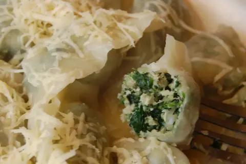 Pyszne pierogi z kurczakiem i szpinakiem, polane sosem i posypane startym serem. Jeden pieróg jest przekrojony, ukazując bogate nadzienie.