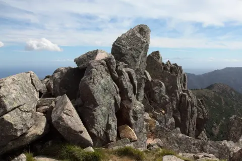 Rochers escarpés au sommet de la Punta di a vacca morta, avec des montagnes en arrière-plan sous un ciel bleu et nuageux.