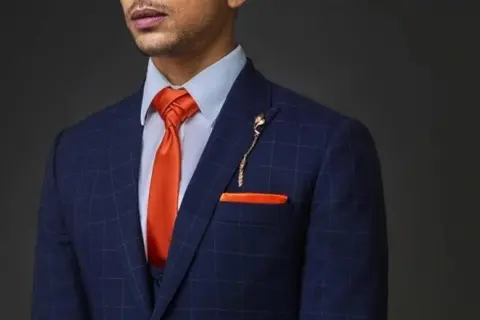Uomo con abito blu a quadri, camicia azzurra e cravatta arancione con nodi cravatta particolari. Taschino e spilla coordinati.