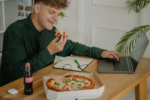 Młody mężczyzna je pizzę, pracując przy laptopie. Obok butelka napoju i notatnik. Dba o dietę, pamiętając o insulinie aspart.