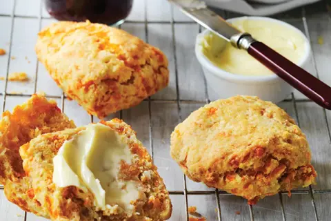 Trois scones américains dorés, dont un coupé en deux et garni de beurre, sur une grille. Un pot de confiture et du beurre à tartiner sont à proximité.