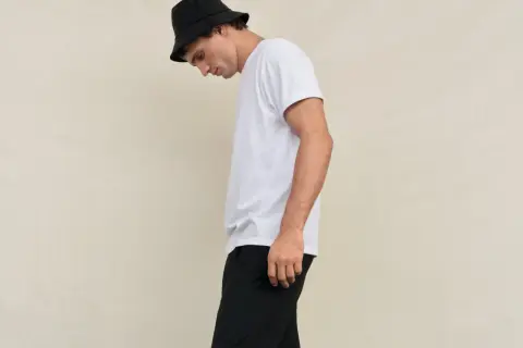 Młody mężczyzna w czarnym kapeluszu typu bucket hat i białej koszulce, spodnie w stylu skate.
