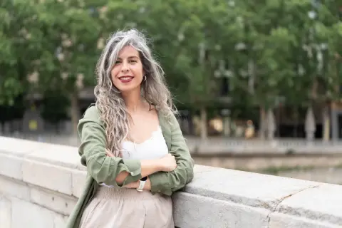 Mujeres pelo rizado con canas, sonriendo con los brazos cruzados, apoyada en un muro de piedra.