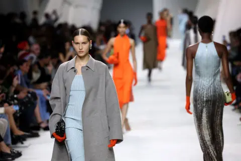Angelina Kendall sfila con un cappotto grigio oversize e un abito a righe azzurre, guanti arancioni vivaci.