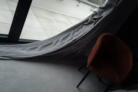 Szare zasłony swobodnie opadają na podłogę, tworząc przytulną atmosferę wokół pomarańczowego fotela.