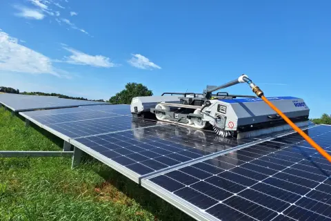 Robot do mycia paneli fotowoltaicznych czyści rzędy paneli na zielonej łące pod błękitnym niebem.