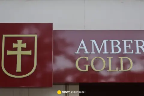 Złoty symbol krzyża na bordowym tle obok napisu AMBER GOLD.