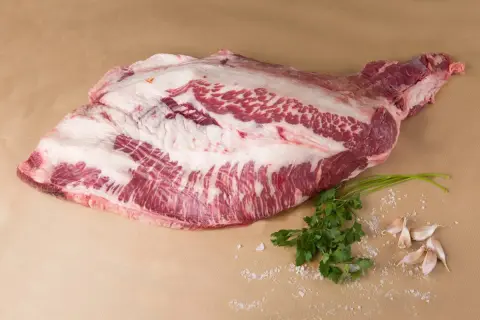 Un corte de carne cruda, con grasa marmoleada, listo para convertirse en un delicioso pastrami que es. Acompañado de perejil y ajos.