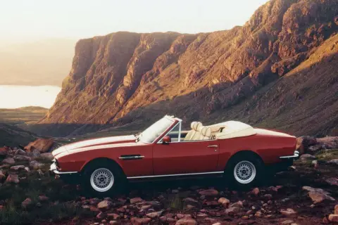Röd Aston Martin V8 Vantage cabriolet parkerad i en stenig, bergig terräng med en sjö i bakgrunden.