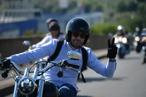 Mężczyzna w kasku i niebieskiej koszuli jedzie na motocyklu i pokazuje znak pokoju.