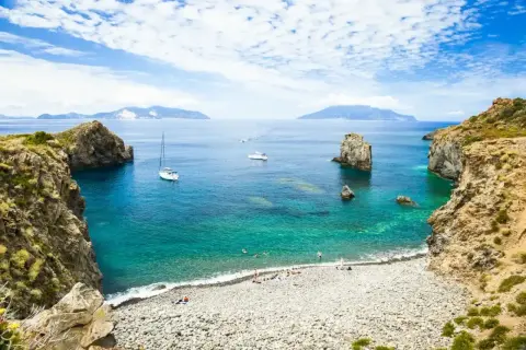 Spiaggia di ciottoli e acque cristalline, ideale per una vacanza alle Eolie. Barche a vela e isole all'orizzonte.