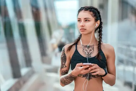 Kobieta z warkoczami i słuchawkami na uszach, trzymająca telefon. Widoczny delikatny tatuaż między piersiami w kształcie kota.
