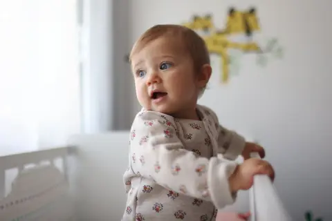 Un bébé aux yeux bleus, prénom fille, se tient dans son lit, curieux du monde.