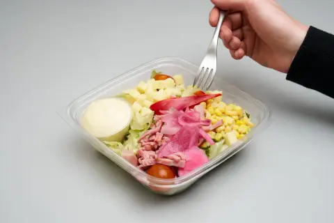 En hand tar en gaffel ur en plastlåda med pastasallad med skinka och fetaost, majs, tomat och dressing.