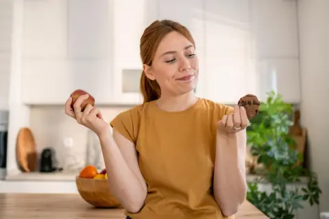 Vrouw twijfelt tussen een appel en een chocoladekoekje. De strijd tegen het verslaafd zijn aan eten is zichtbaar in haar blik.
