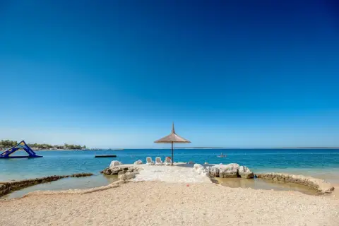 Słoneczna plaża z parasolem i leżakami w camping village Simuni. W tle wodny plac zabaw i spokojne morze.