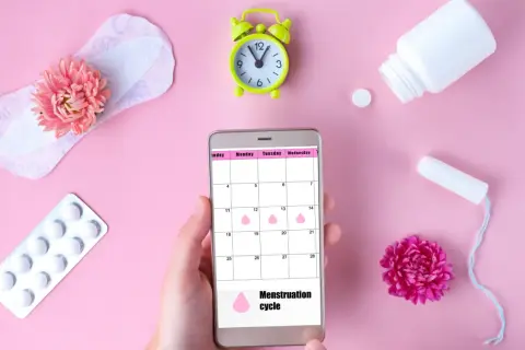 Telefon z kalendarzem cyklu menstruacyjnego, obok podpaska, tampon, leki i kwiaty. Warto pamiętać o mięśniakach macicy.