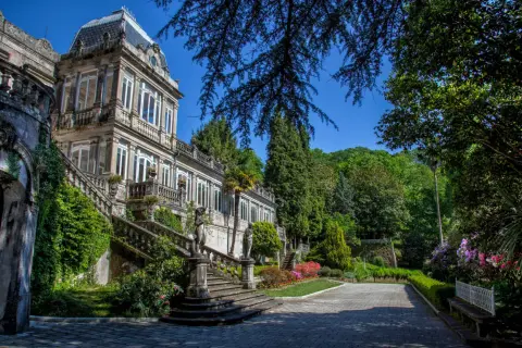 El Pazo de Lourizán, una mansión señorial con escalinatas cubiertas de hiedra y un jardín exuberante, se alza majestuoso bajo un cielo azul.