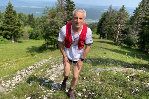 Un coureur en tenue de trail, sac à dos rouge, sur un sentier escarpé près d'Oyonnax, profitant d'un paysage verdoyant et vallonné.