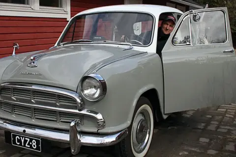 Man ler vid ratten i en grå Morris Oxford med öppen dörr.