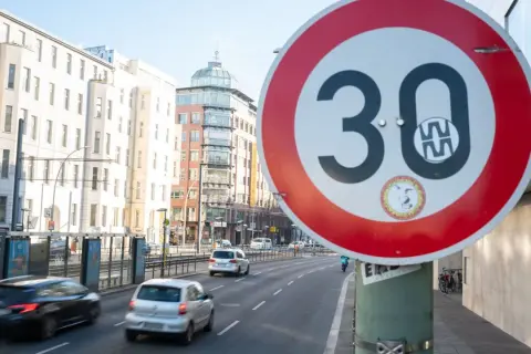 Verkehrsschild "Tempo 30 Berlin" auf einer Straße mit Autos und Gebäuden im Hintergrund.
