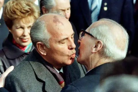 Gorbatschow und Honecker im Bruderkuss. Eine Frau im Hintergrund lächelt.