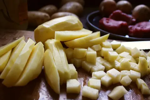 Skalade och skurna potatisar i olika former, redo för att bli härligt råstekt potatis. I bakgrunden syns hela potatisar och kött.