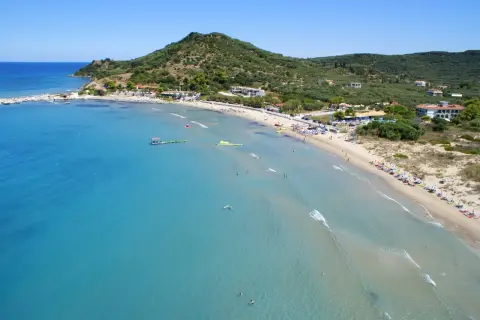 Słoneczny dzień na plaży Alykanas. Turkusowa woda, piaszczysty brzeg z leżakami i parasolami, zielone wzgórza w tle.