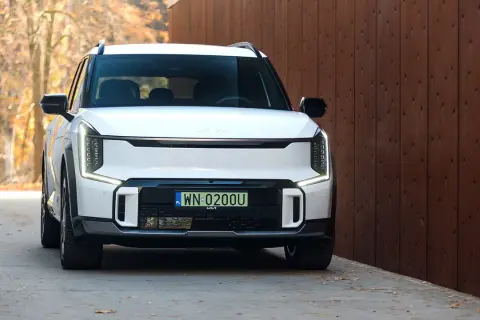 Biały Kia EV9 z polskimi tablicami WN 0200U zaparkowany przy drewnianym ogrodzeniu, jesienne liście na ziemi.
