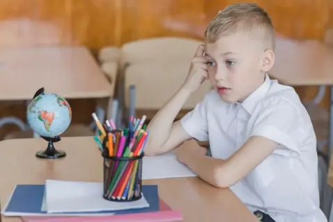 Chłopiec w białej koszuli siedzi przy biurku z globusem i kredkami. Zamyślony, może czeka na odroczenie obowiązku szkolnego.