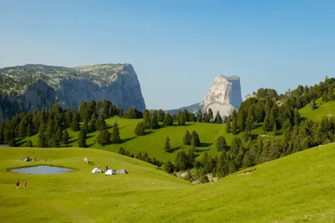 Un campement de tentes et de randonneurs sur une prairie verdoyante, avec des montagnes imposantes en arrière-plan. Le **pas de l'aiguille** est visible au loin.
