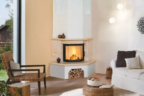 Gemütliches Wohnzimmer mit einem Eck-Kachelofen, der einladend brennt. Ein Rattansessel und ein weißes Sofa laden zum Entspannen ein.