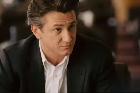 Sean Penn w czarnym garniturze i białej koszuli, wygląda jakby zastanawiał się nad kolejną rolą w swoich filmach.