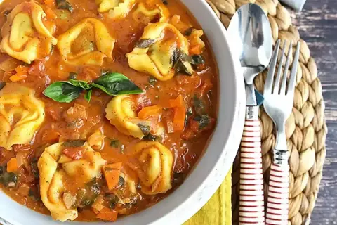 Pyszne tortellini z mięsem w kremowym sosie pomidorowym z warzywami, udekorowane listkiem bazylii. Obok widelec i łyżka.